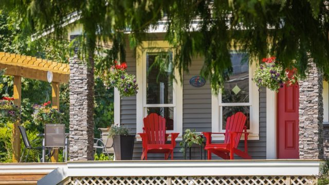 View of a large front porch with furniture and potted plants. Rustic, country style front porch seating with red Adirondack chairs and fresh flowers.