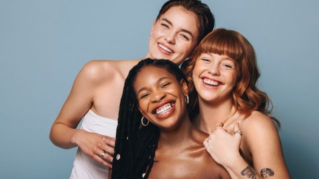 Cheerful women with different skin tones smiling at the camera in a studio. Group of happy young women embracing their natural skin. Portrait of three body positive young women standing together.