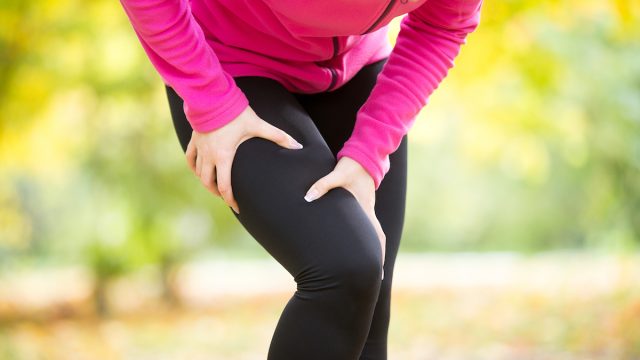 woman wearing black leggings and a pink top grabbing her leg in pain while exercising outside