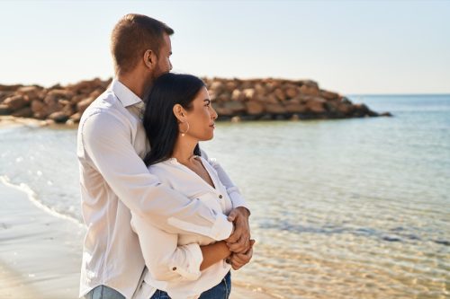 Man and woman couple smiling happy hugging each other standing at seaside