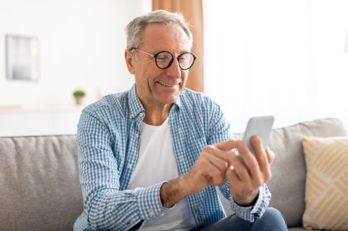 Portrait Of Cheerful Mature Guy In Glasses Using Mobile Phone With New Application Sitting On Sofa In Living Room. Gadgets And Mobile Communication, Older People And Technologies Concept