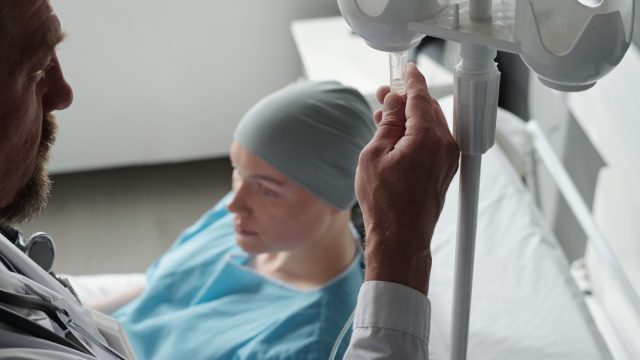 Close-up of mature male oncologist preparing dropper for chemotherapy before procedure while standing by bed with sick female patient