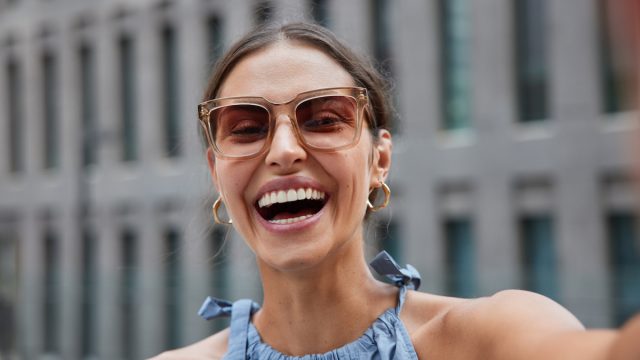 portrait of a happy, laughing woman taking a selfie while outside on a city street wearing oversized sunglasses