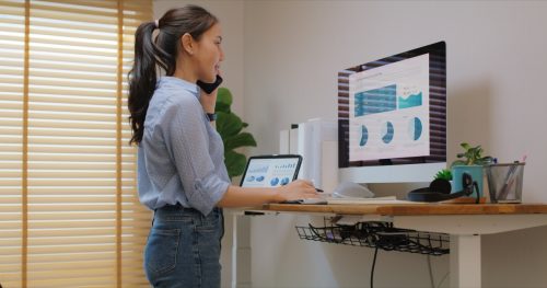 Electric adjustable standing desk in small office workspace for PC desktop computer worker. Asia people young woman relax remote work at home proper height up workstation physical workforce challenge.