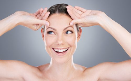 woman doing a face yoga exercise, raising her eyebrows, against a gray background