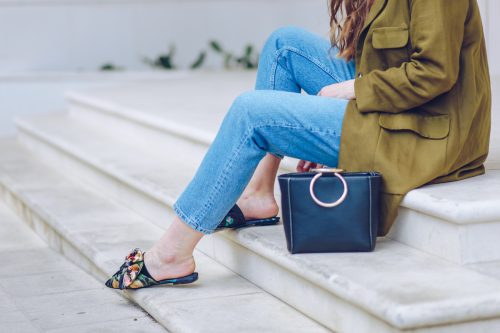 cropped image of a woman wearing jeans and floral mules with an olive green jacket while sitting on outside steps