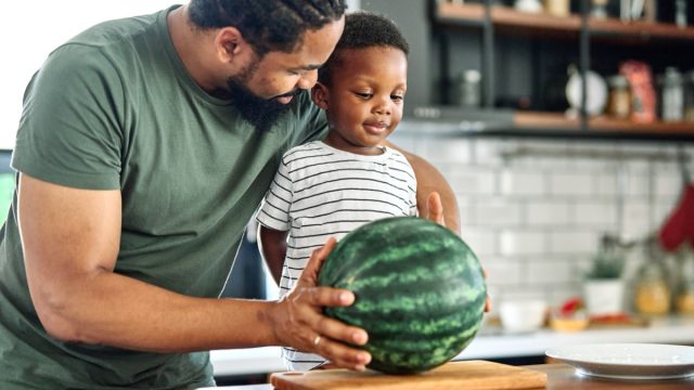 Man and little boy standing at kitchen counter with whole watermelon on cutting board