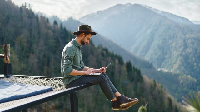 Young man freelancer traveler wearing hat anywhere working online using laptop and enjoying mountains view