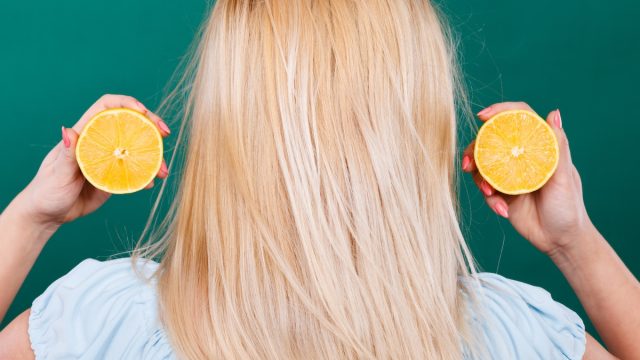 back of a blonde woman holding two halfs of a lemon on a dark green background