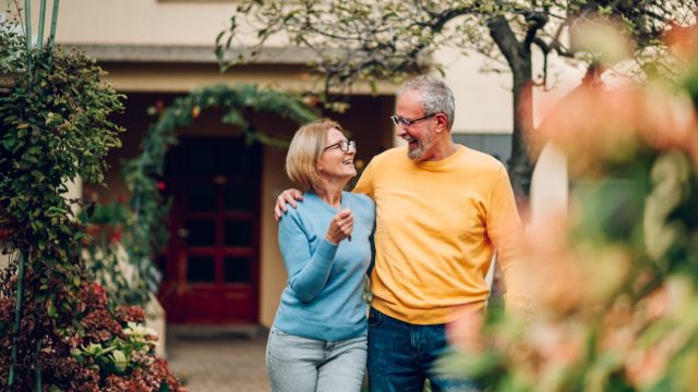 Happy mature couple bought their new home and standing in front of it and showing keys. Real estate owners. Senior married couple posing in front of their new family house. Looking at each other.