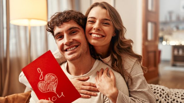 A woman gives a greeting card to her beloved man with the inscription I love you in the living room at home.