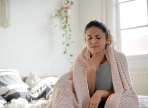 woman sitting up in bed with a blanket wrapped around her, holding her throat to indicate a sore throat