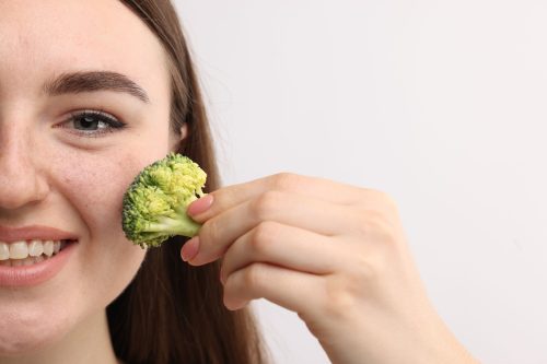 Smiling woman making fake freckles with broccoli and cosmetic product on light background, closeup.